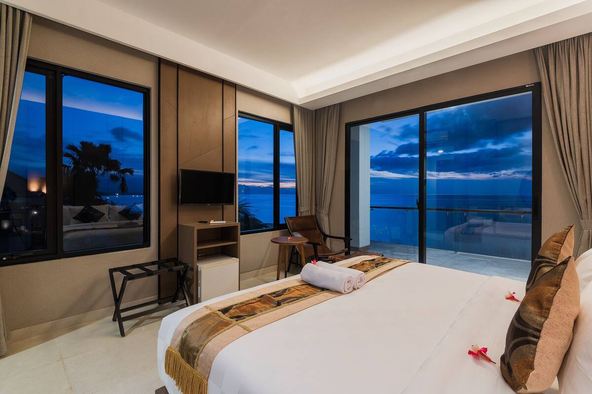 A hotel room with a large bed, a desk, a chair, a TV, and large windows with a view of the ocean.