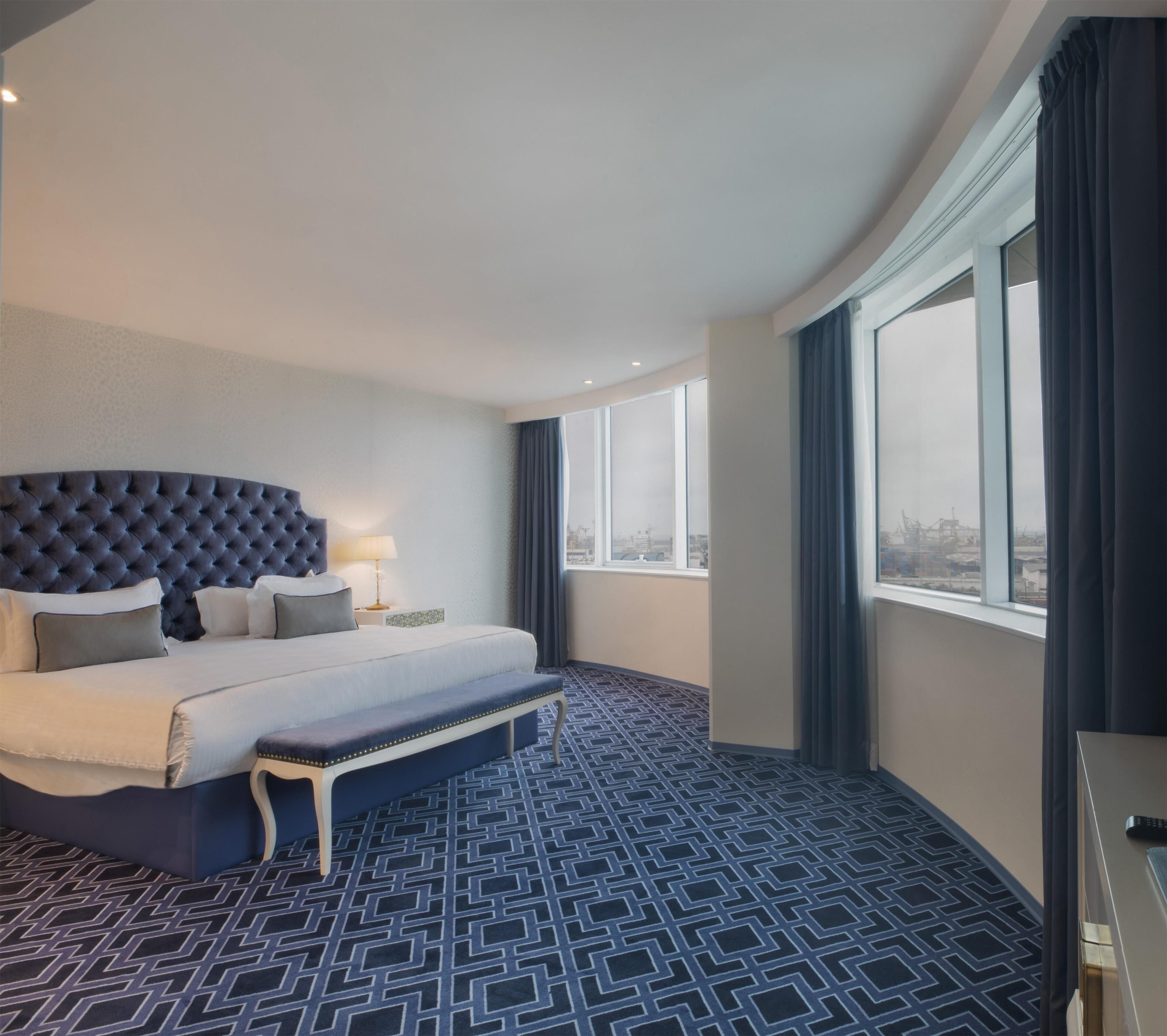 Ein Hotelzimmer mit einem großen Bett, einer Sitzbank, einem Nachttisch und Blick auf die Stadt durch große Fenster.