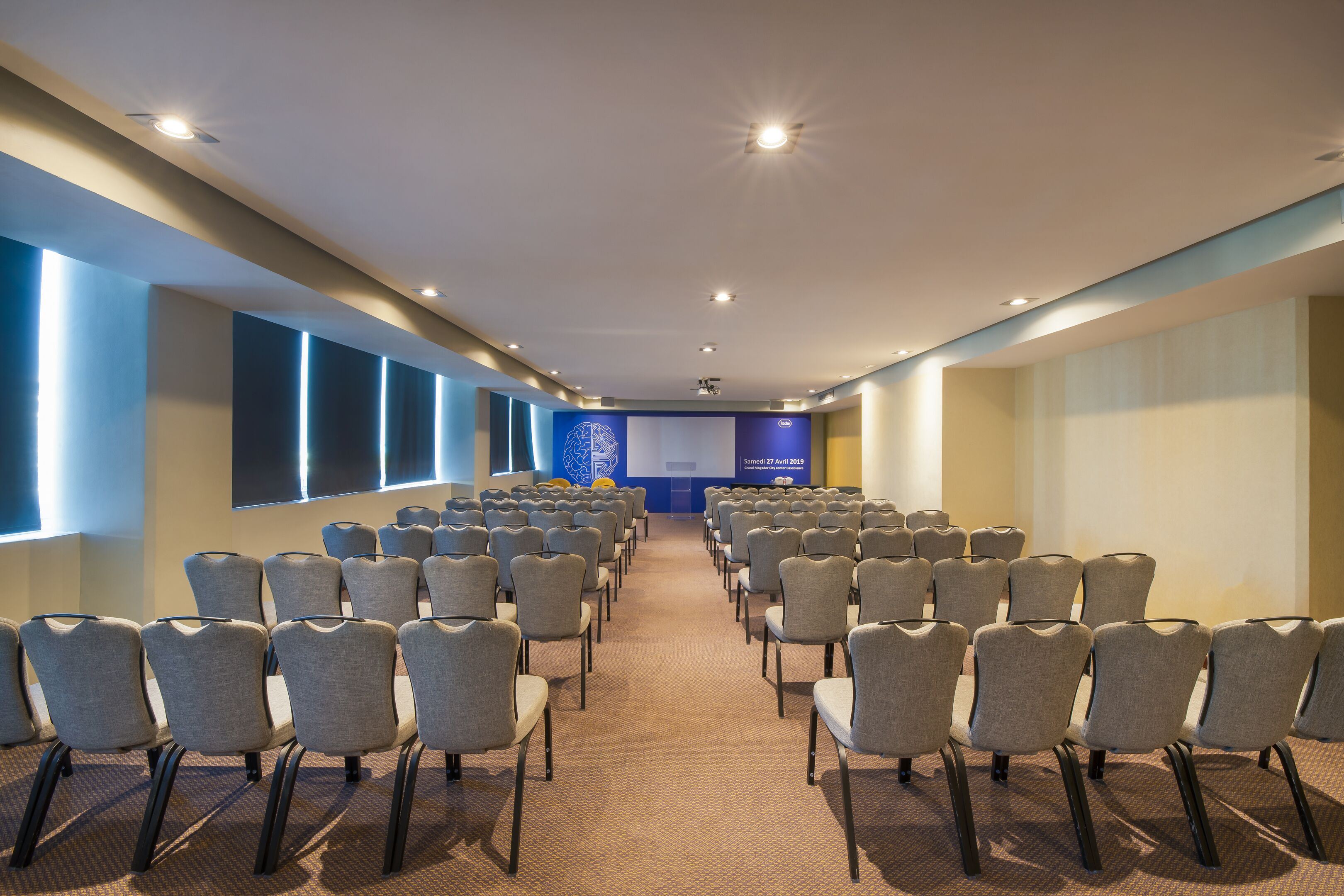 A conference room with rows of chairs facing a presentation screen, carpeted floor, and recessed ceiling lights.
