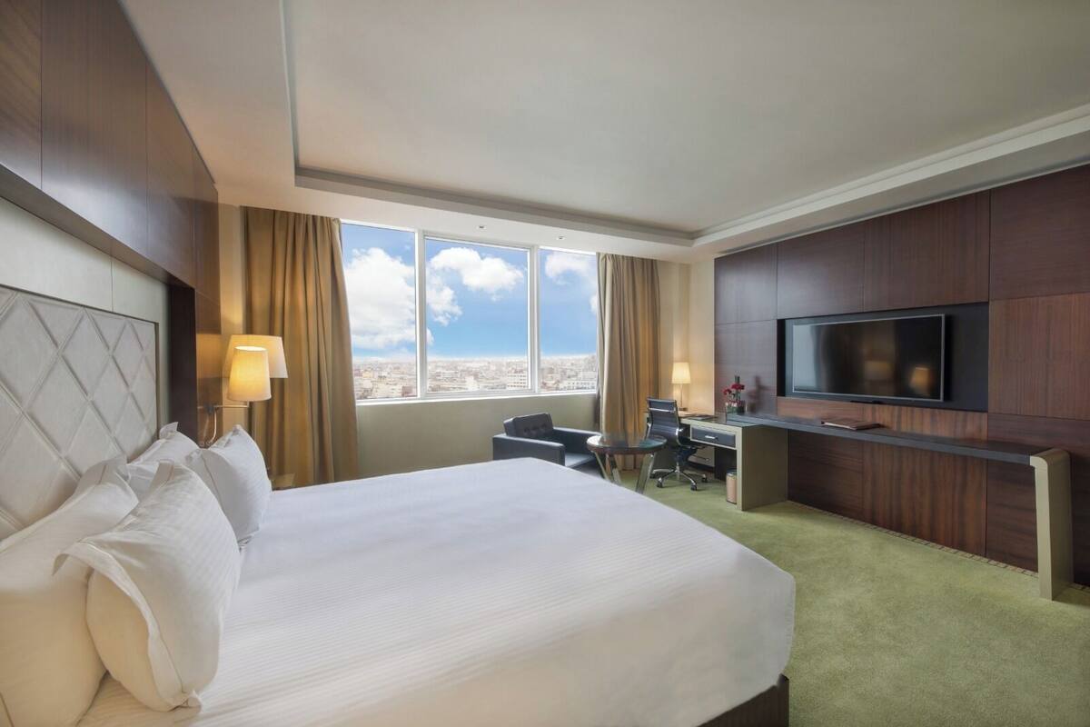 Une chambre d’hôtel dotée d’un grand lit, d’un bureau, d’une chaise, d’une télévision et offrant une vue sur la ville grâce à de grandes fenêtres.