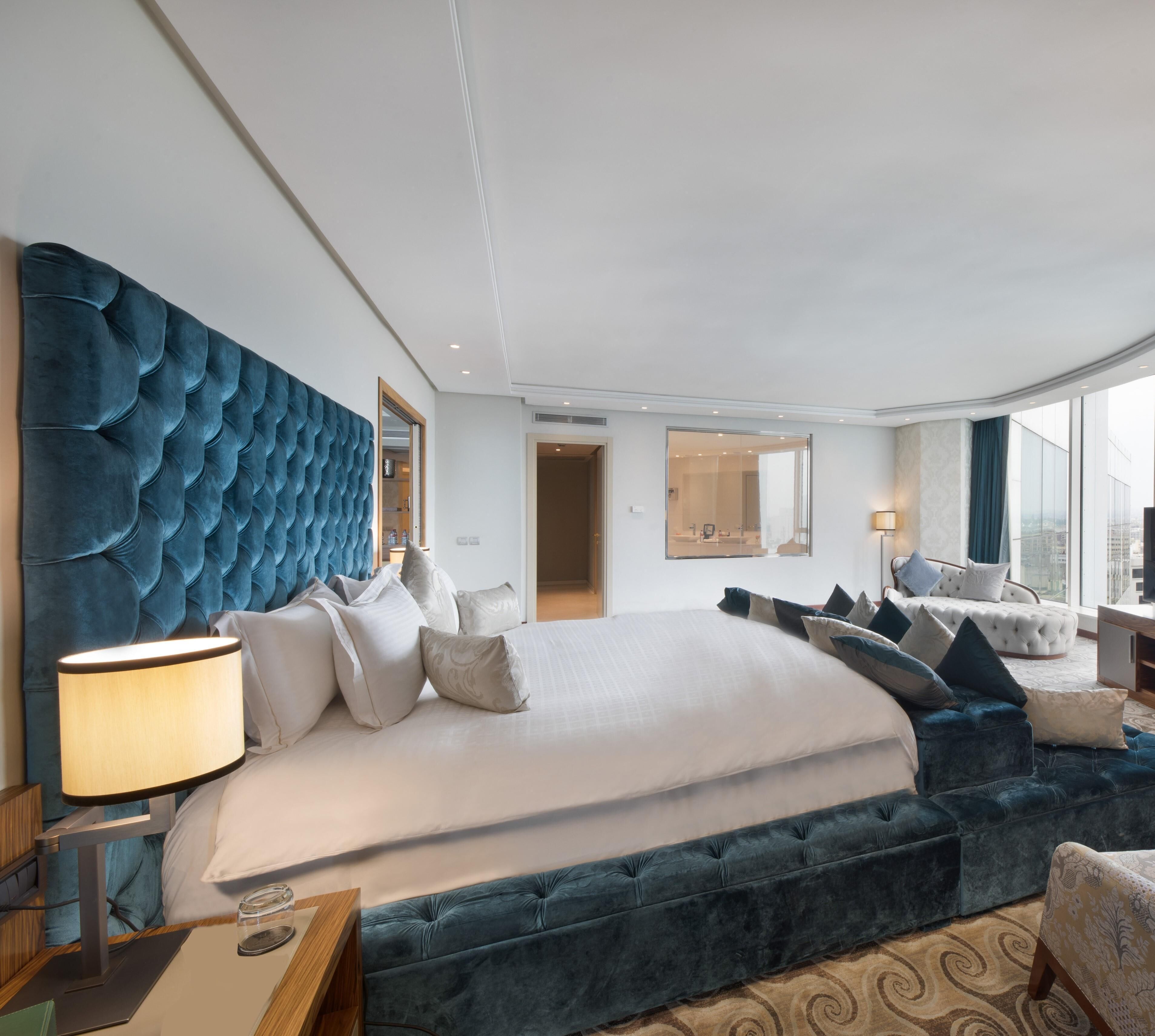Una habitación de hotel moderna con una cama grande, un cabecero capitonado, una zona de estar con sofá y vistas a la ciudad a través de amplias ventanas.