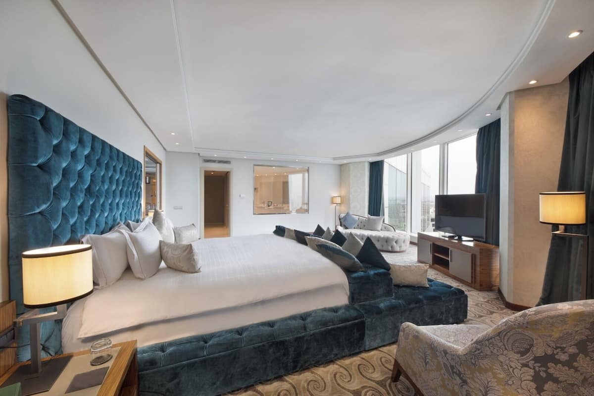 Une chambre d’hôtel moderne dotée d’un grand lit, d’un coin salon avec des canapés bleus, d’un téléviseur à écran plat et d’une grande fenêtre offrant une vue sur la ville.