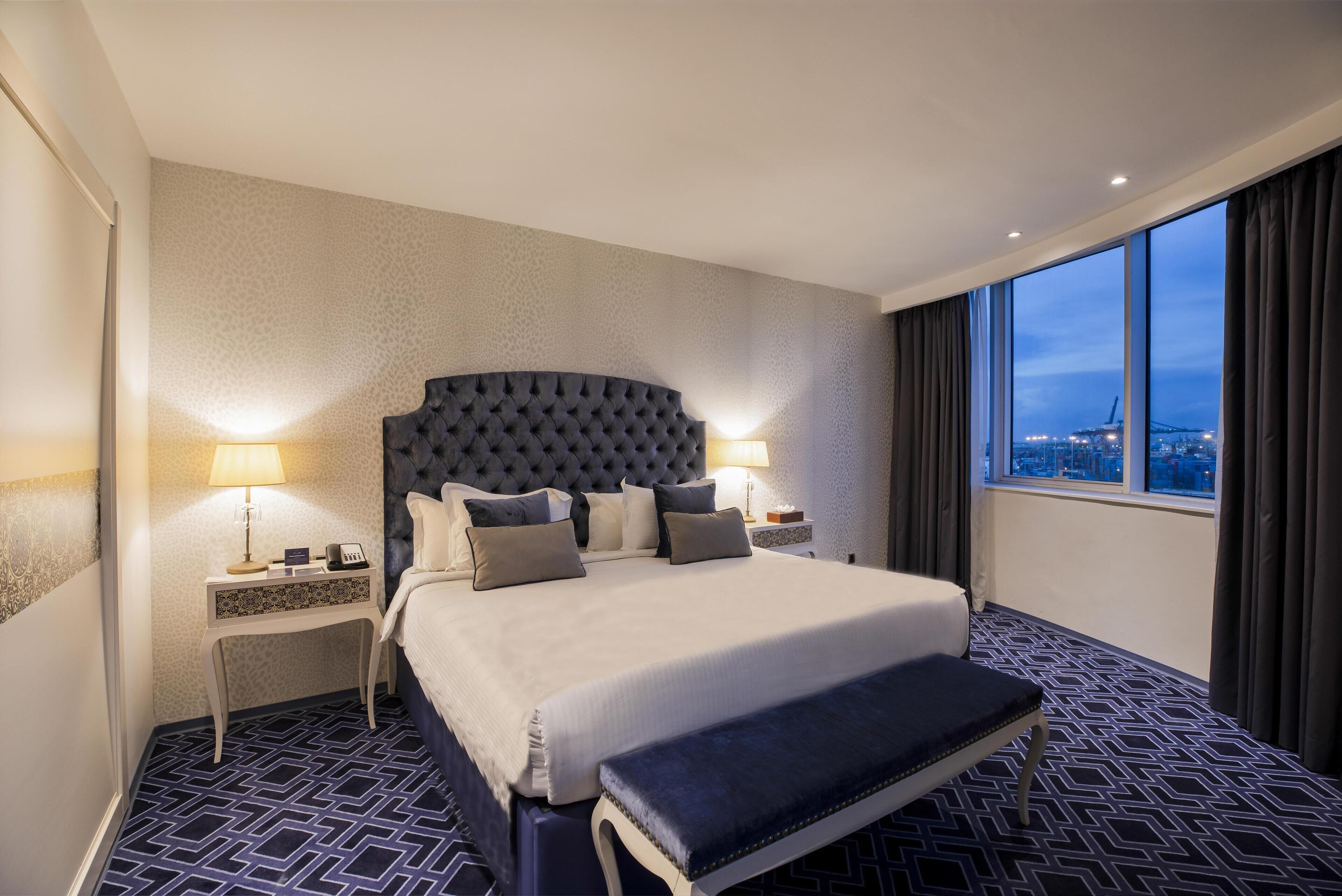 Ett hotellrum med en stor säng, två sängbord med lampor, en bänk och utsikt över staden genom stora fönster.