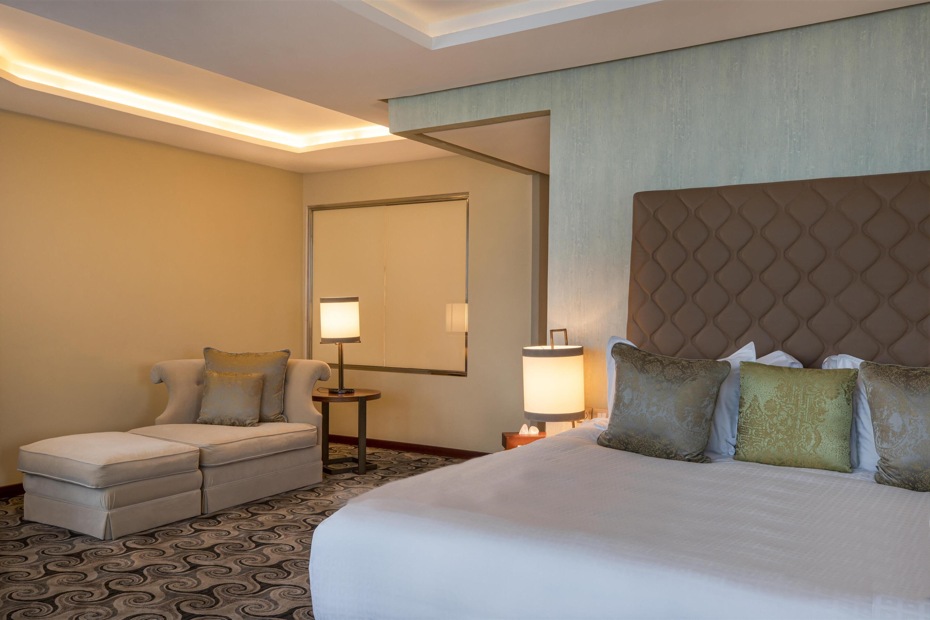 A hotel room with a large bed, a sofa, two bedside tables with lamps, and a patterned carpet.
