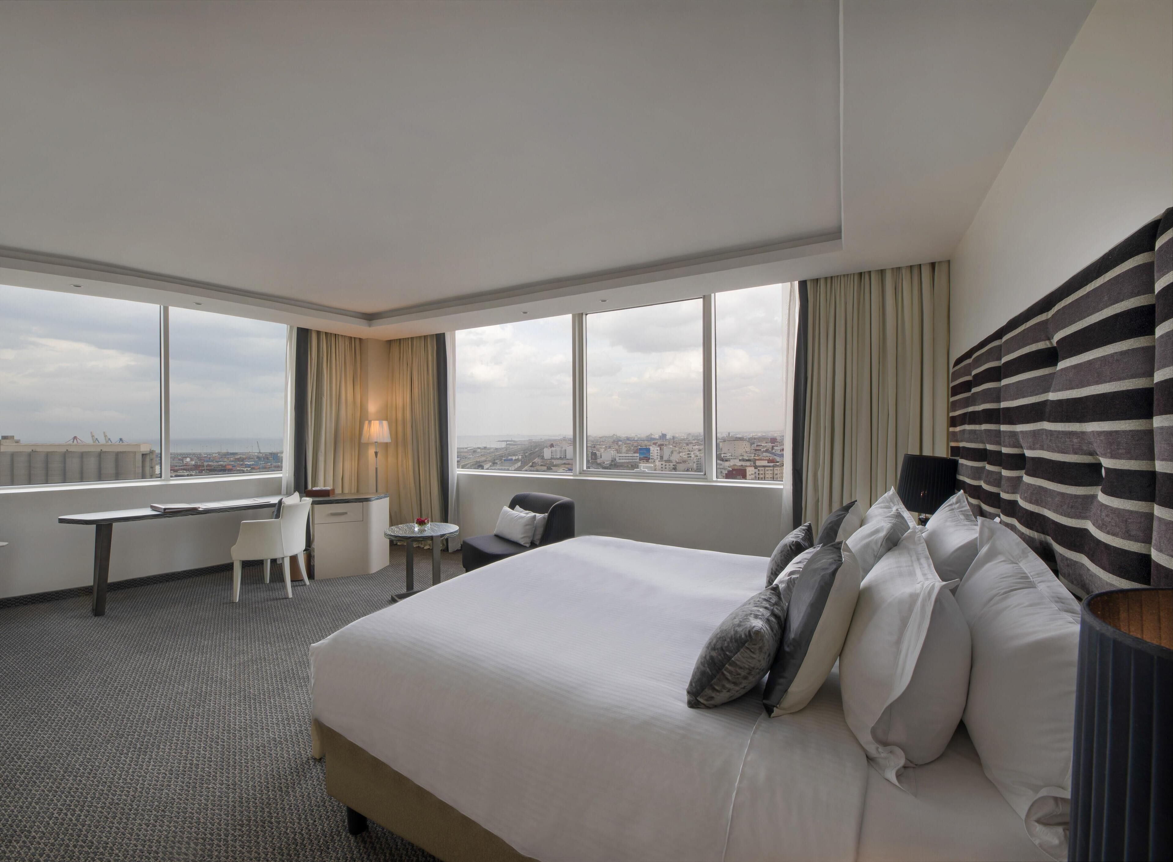 Une chambre d’hôtel avec un grand lit, un bureau, une chaise et une vue sur le paysage urbain grâce à de grandes fenêtres.