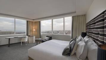 Habitación de hotel con una cama grande, un escritorio, una silla y vistas de la ciudad a través de amplios ventanales.