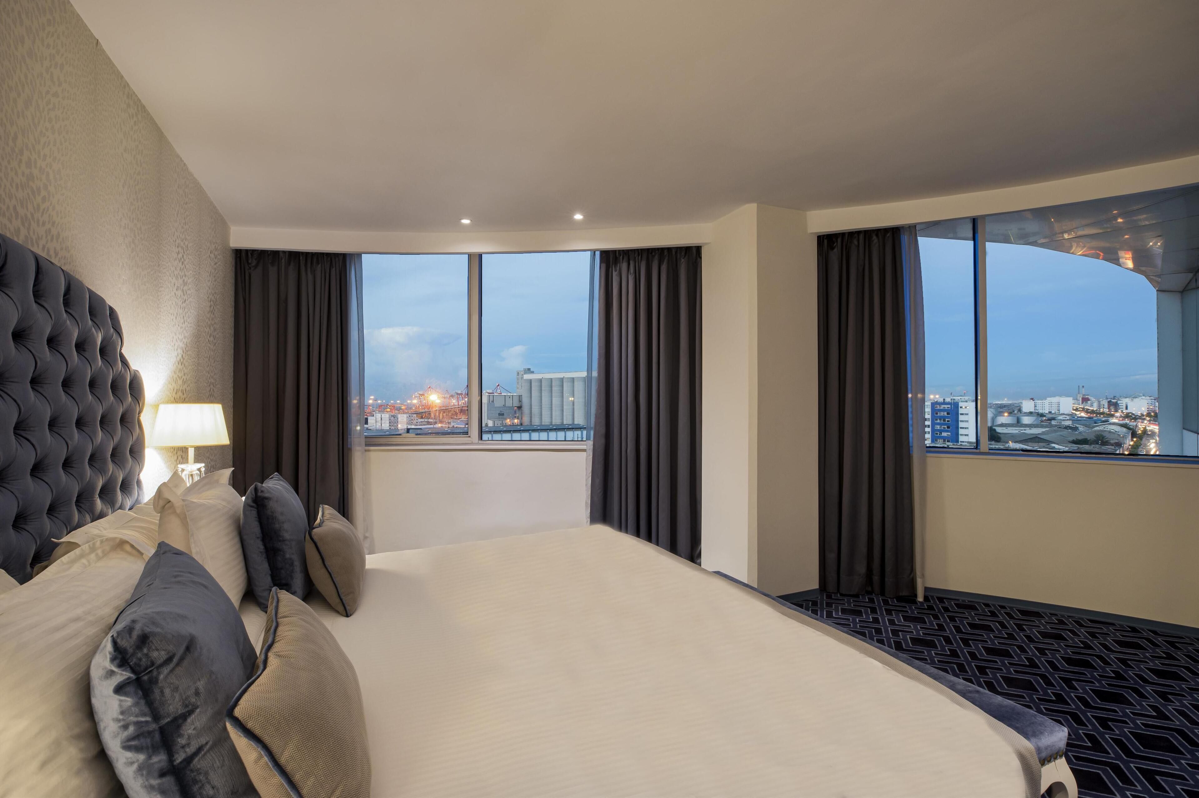 A hotel room with a large bed, a tufted headboard, a view of the cityscape, and a large window with curtains.