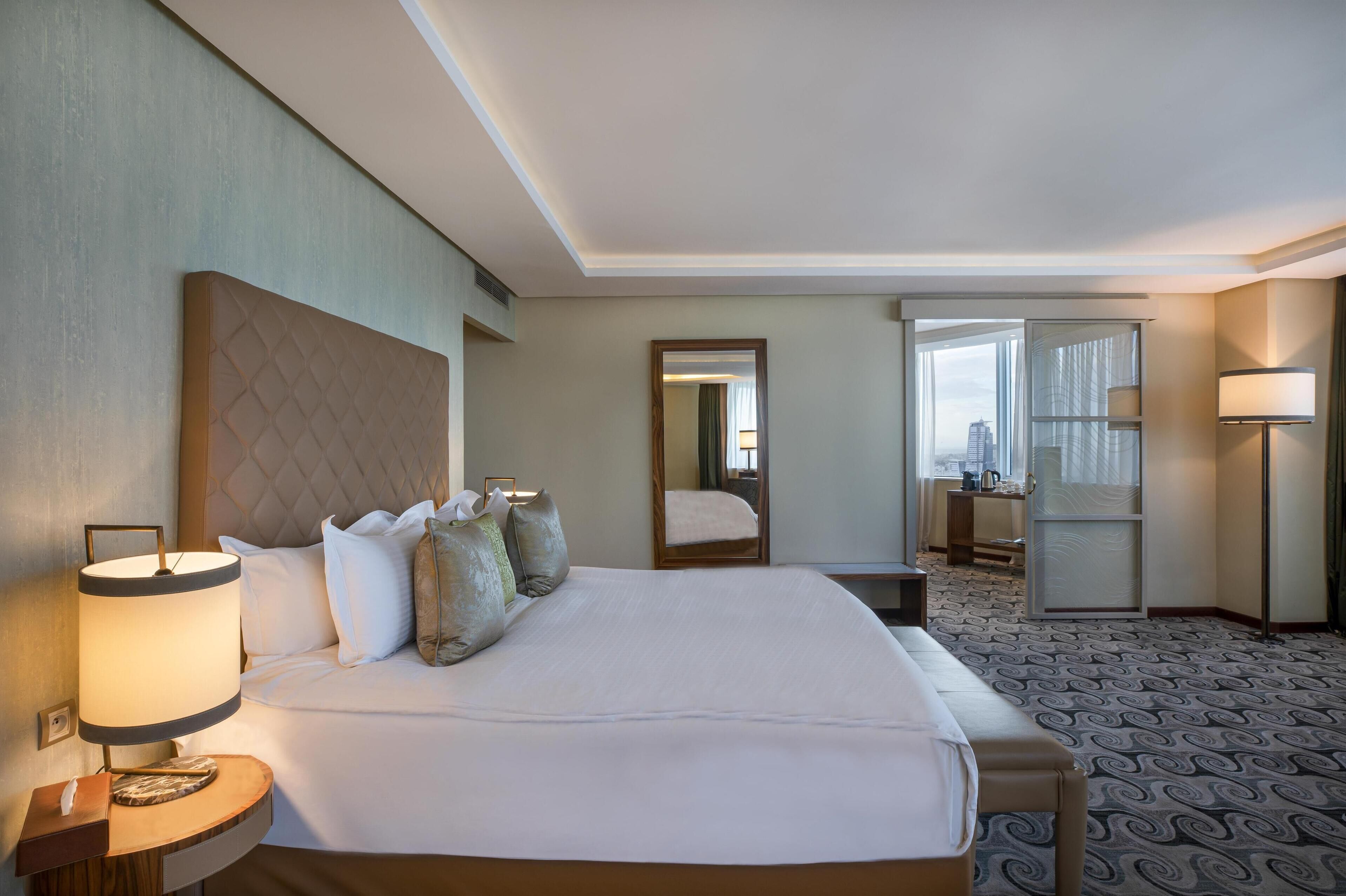 A hotel room with a large bed, a bedside table with a lamp, a mirror, and a view of the city through the window.