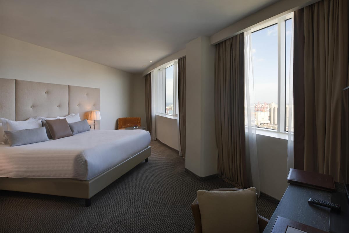 Une chambre d’hôtel avec un grand lit, un bureau, une chaise et une vue sur la ville par les fenêtres.