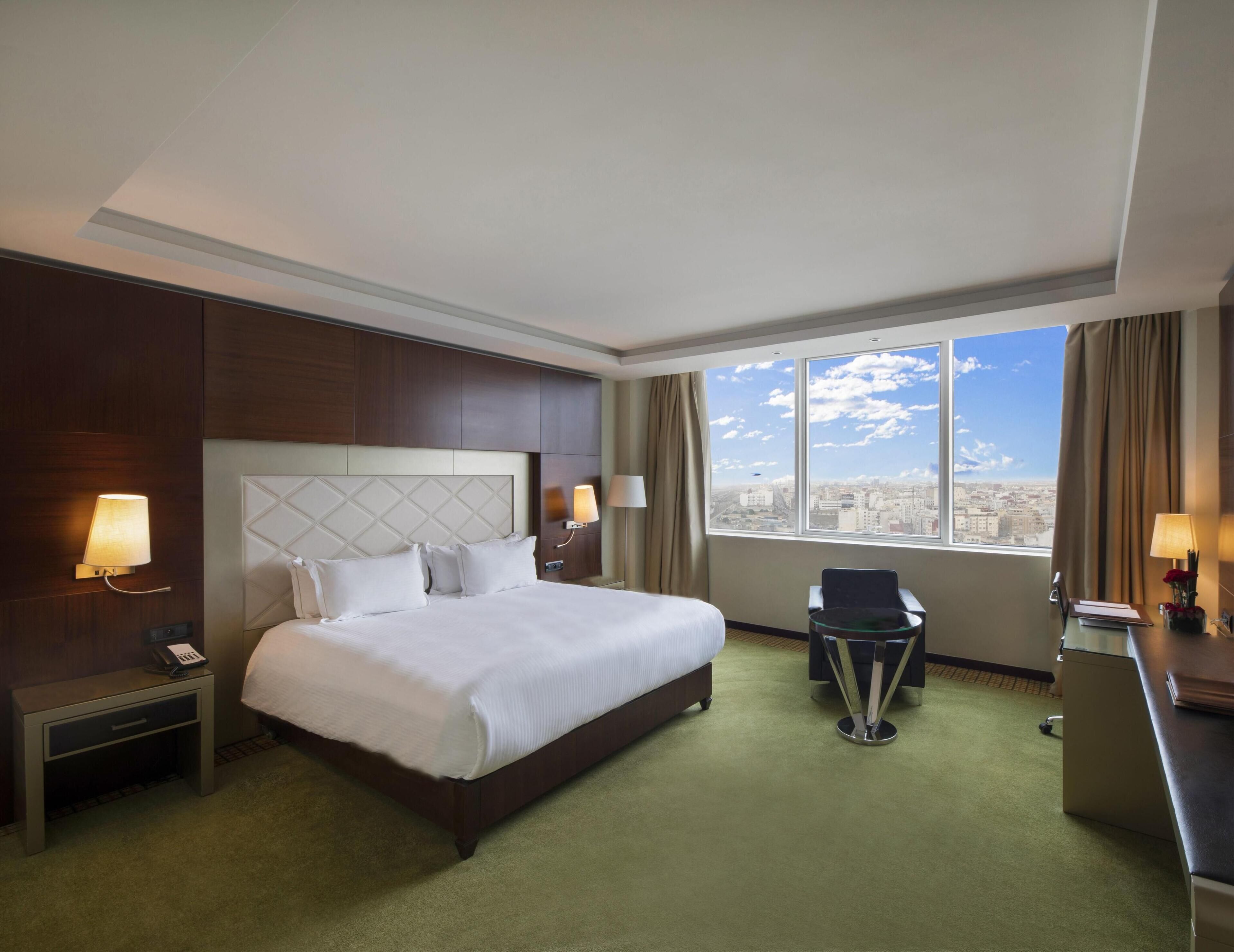 Ein Hotelzimmer mit einem großen Bett, einem Schreibtisch, einem Sessel, einem Telefon auf dem Nachttisch und Blick auf die Stadt durch das Fenster.