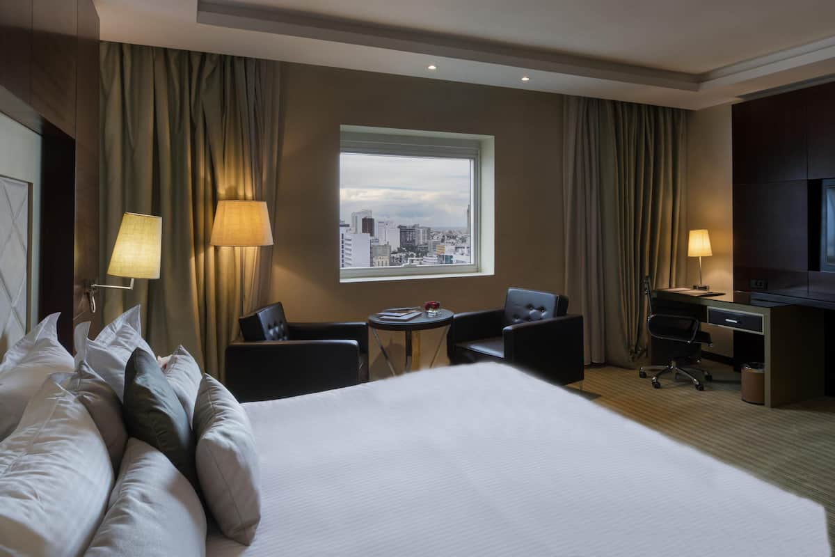 Une chambre d’hôtel avec un grand lit, deux fauteuils, une petite table, un bureau et une fenêtre donnant sur la ville.