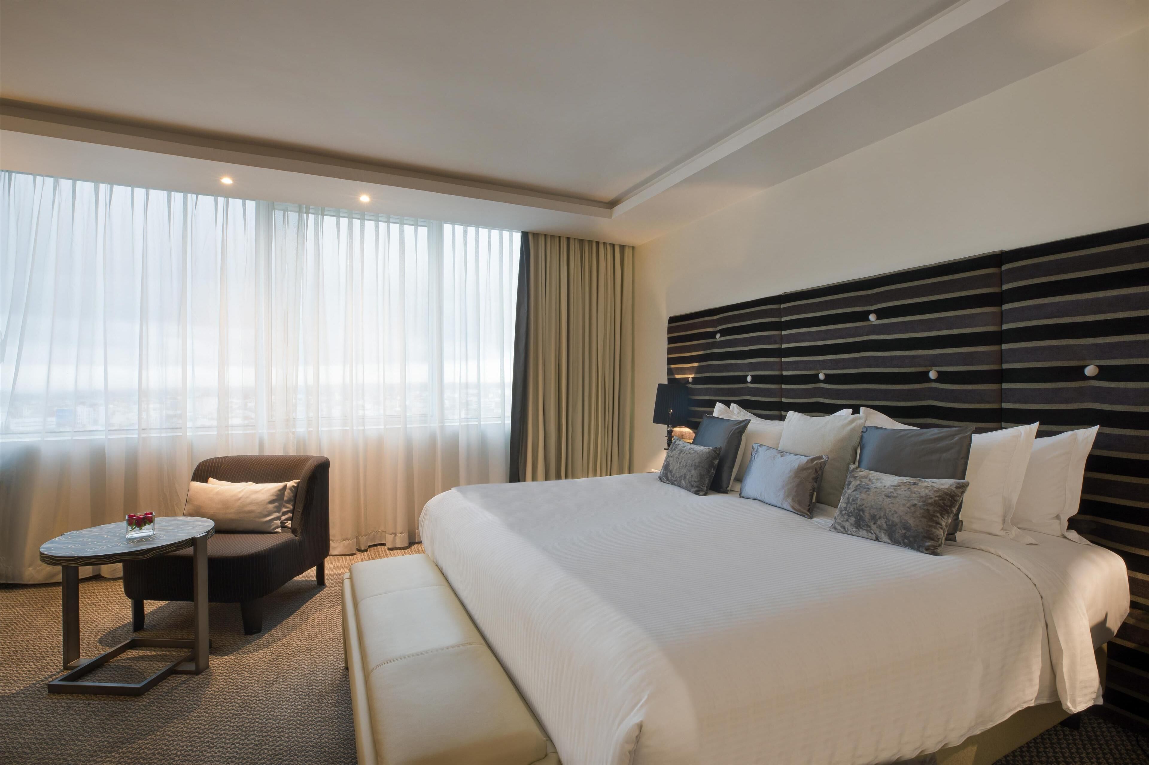 A hotel room with a large bed, a chair, a small table, and a view of the city through sheer curtains.
