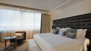 A hotel room with a large bed, a chair, a small table, and a view of the city through sheer curtains.
