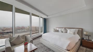 A hotel room with a large bed, a sofa, a small table with a vase of flowers, and a view of the city through large windows.