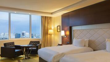 A hotel room with a large bed, two armchairs, a small table, and a city view through large windows.