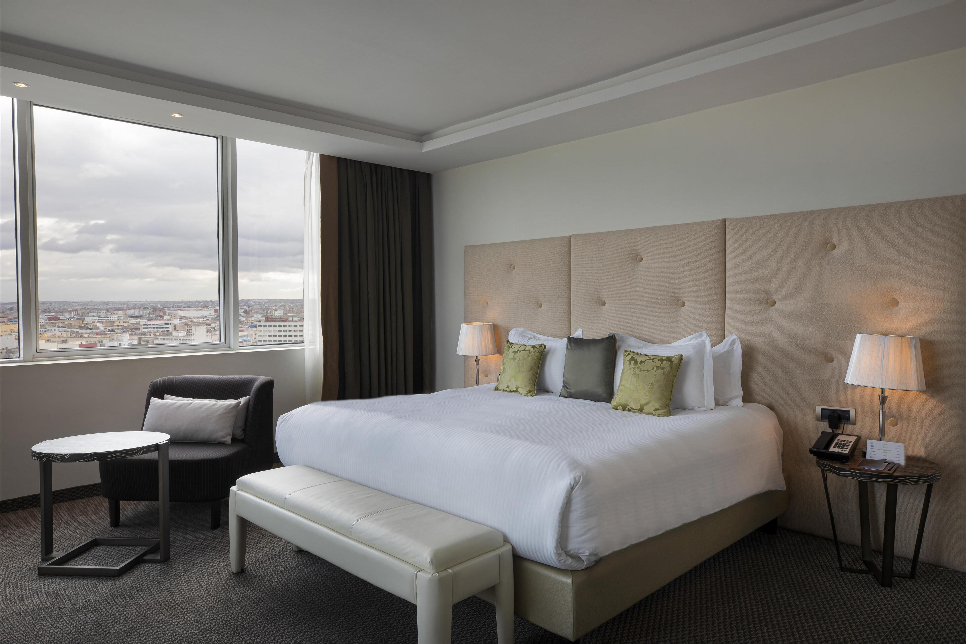 Habitación de hotel con una cama grande, una silla, una mesita pequeña y un banco. Se ve la ciudad a través de amplias ventanas.