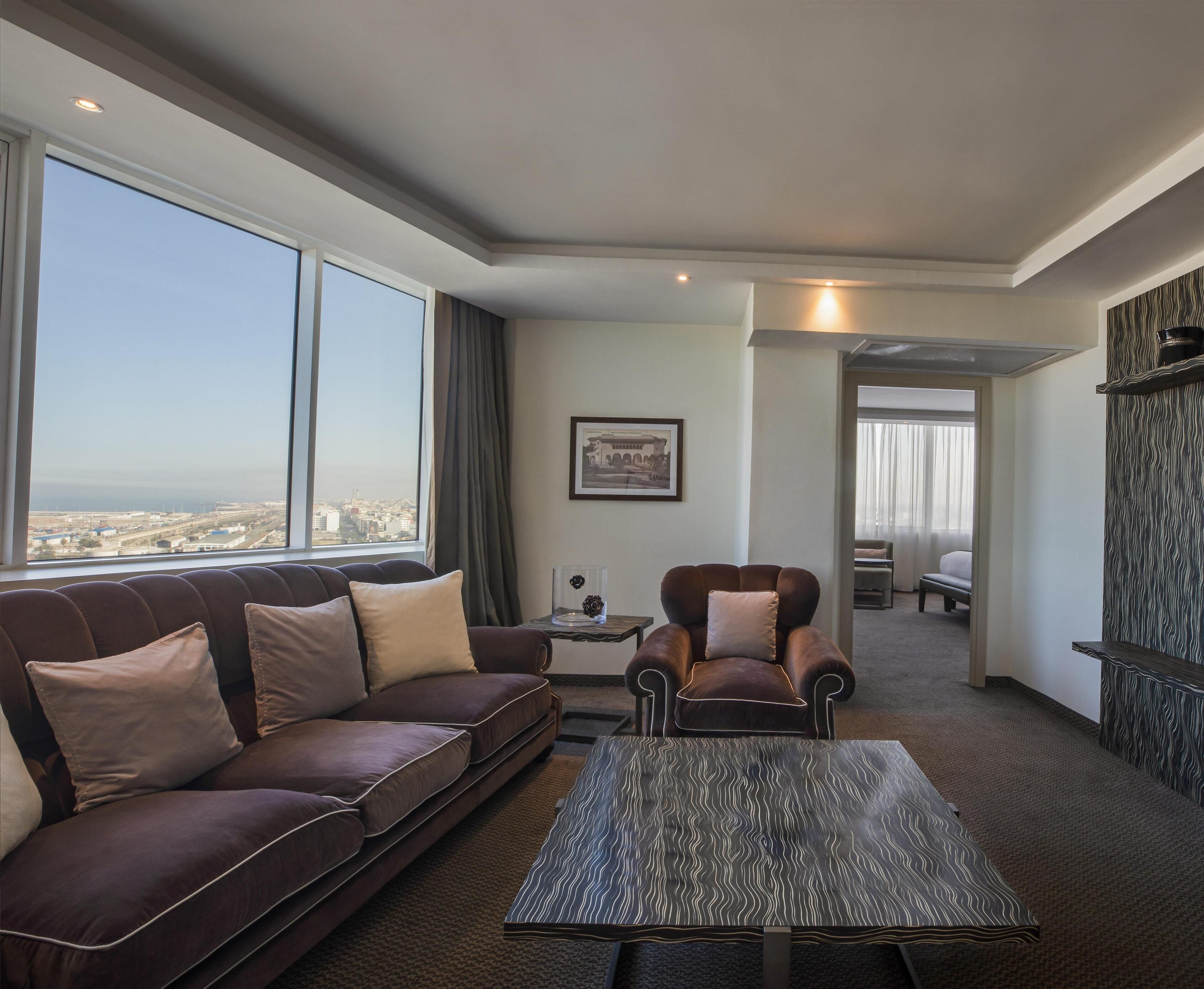 A modern hotel room with a large window offering a city view, a brown leather sofa, a patterned coffee table, and a framed picture on the wall.