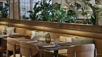 A restaurant interior with a long wooden bench, cushioned chairs, and a variety of plants in the background.