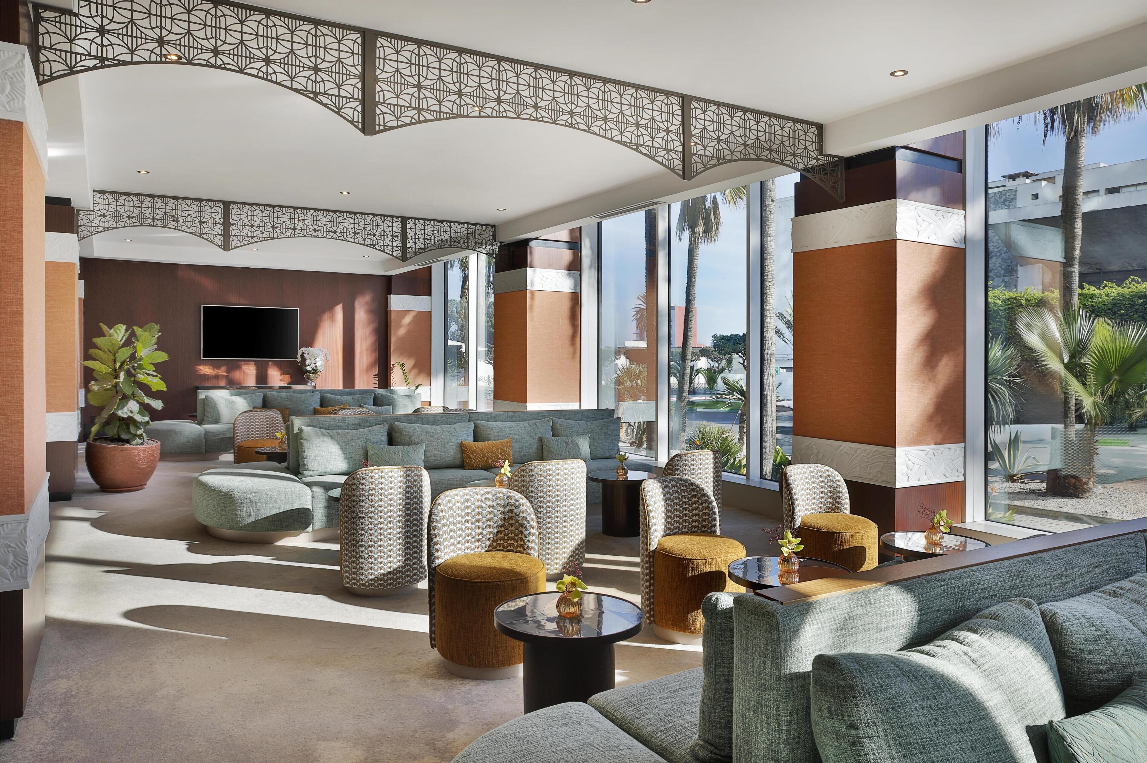 A spacious lobby with a modern design, featuring a seating area with sofas and chairs, a large TV screen, and large windows offering views of palm trees and buildings.
