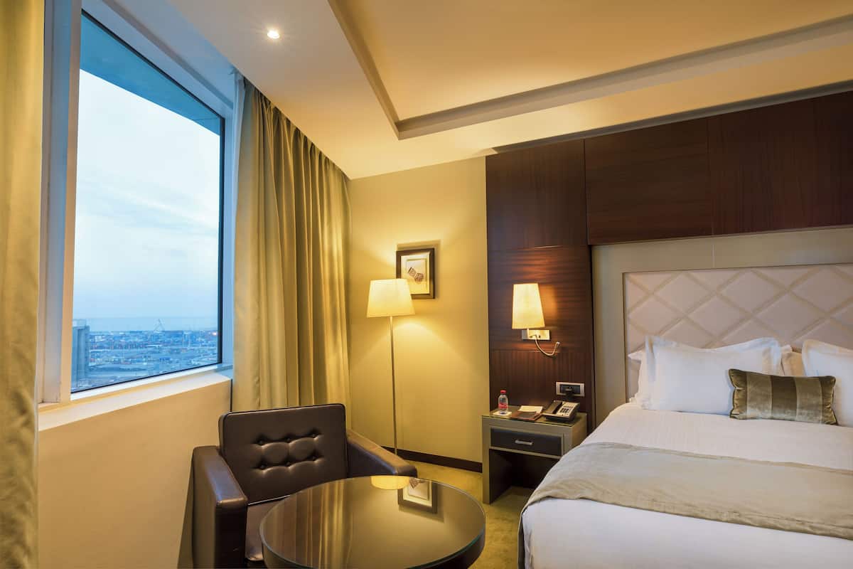 Une chambre d’hôtel équipée d’un lit, d’un bureau, d’une chaise, d’une lampe et offrant une vue sur la ville par une fenêtre.