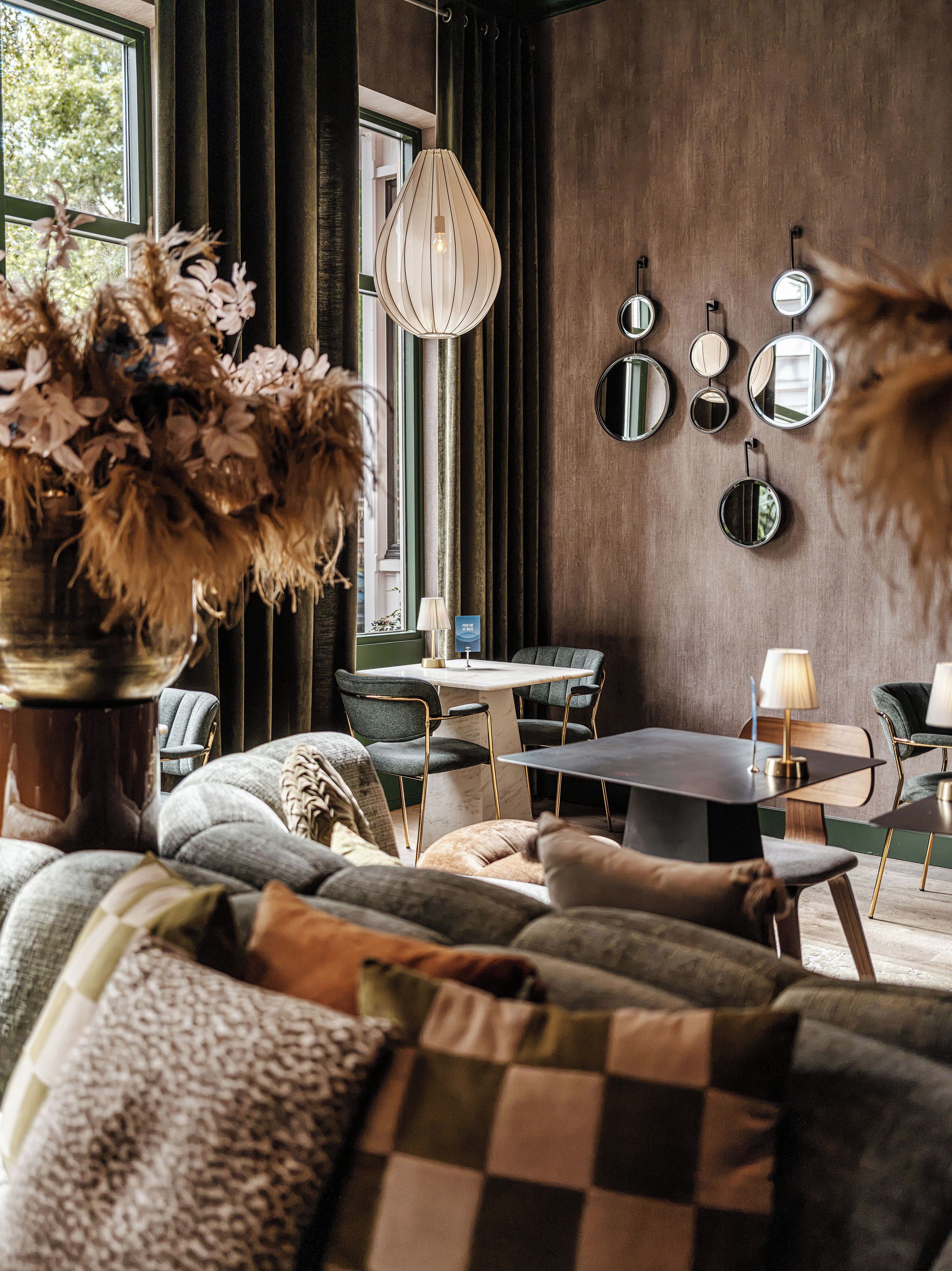 A modern living room with a sofa, a coffee table, and a dining area. There is a vase with dried flowers on a table, and a wall-mounted mirror collection.