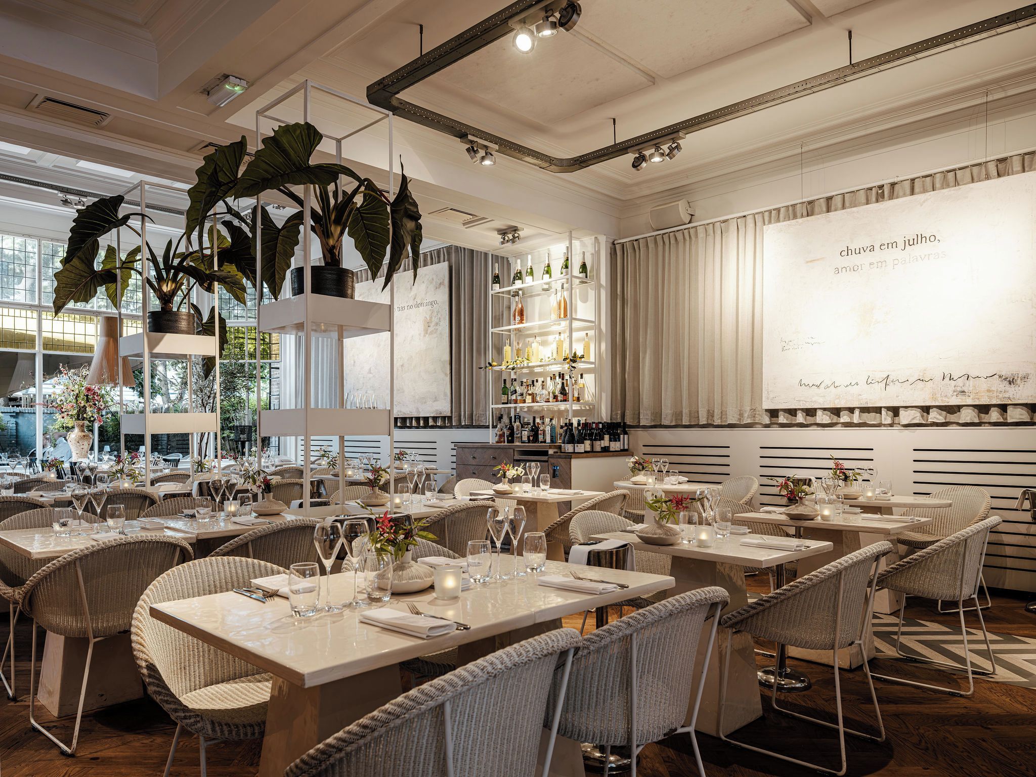 Ein Restaurantinneres mit Esstischen, Korbstühlen und einer Bar mit Flaschenregalen.