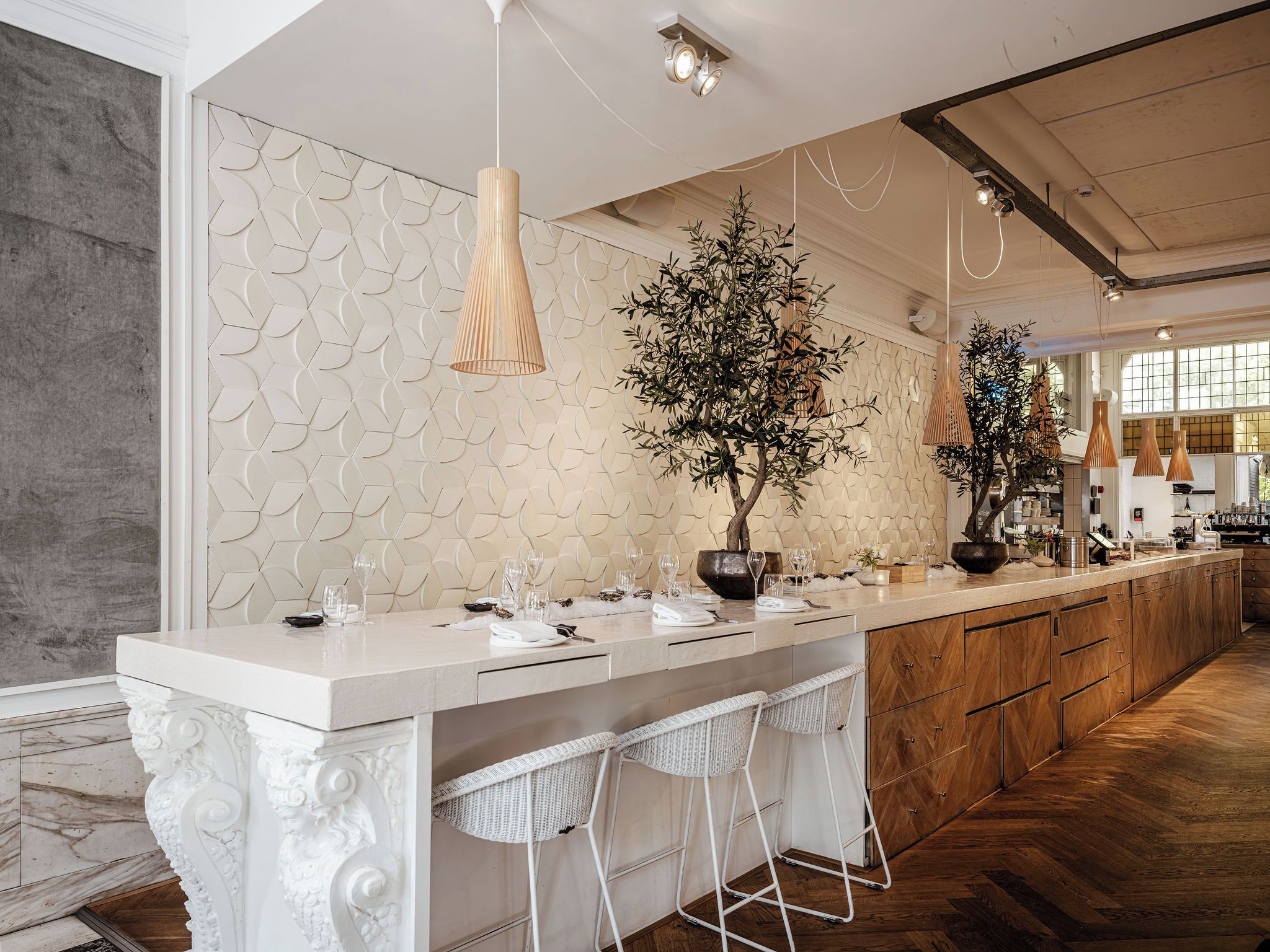 Une cuisine moderne avec un plan de travail blanc, des tabourets de bar, des luminaires suspendus et un mur décoratif.