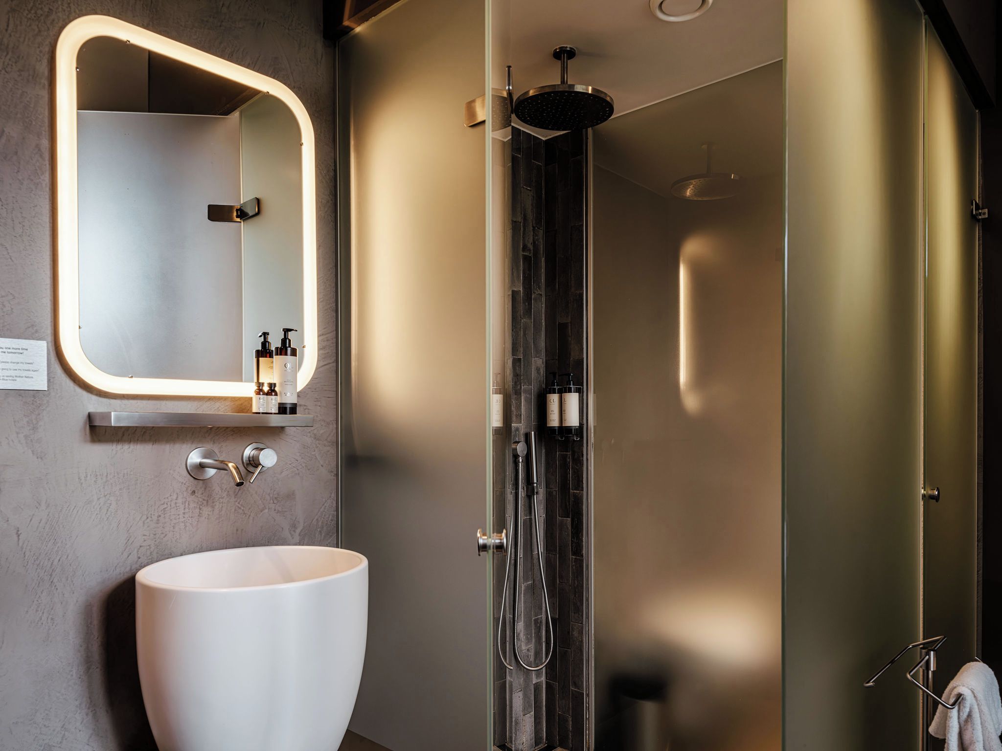 A modern bathroom with a large white sink, a mirror with a light, and a shower area.