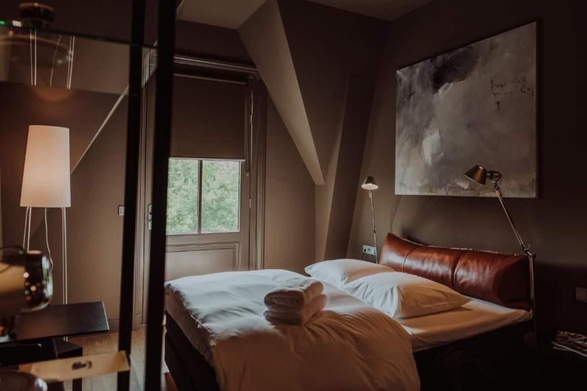 Un dormitorio con cama, escritorio, lámpara, ventana y un cuadro en la pared.