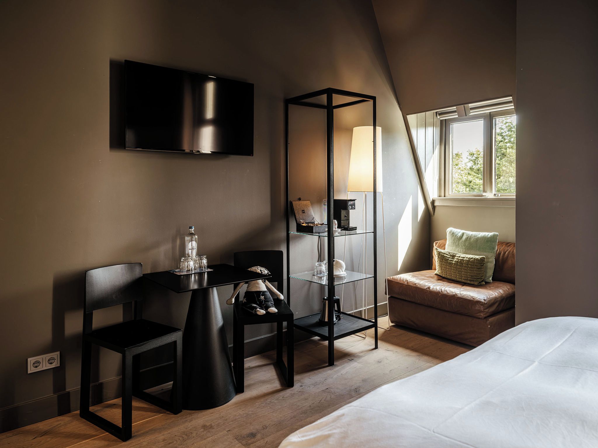 A modern hotel room with a flat-screen TV, a glass table with a bottle and glasses, a black chair, a sofa with cushions, and a window with natural light.