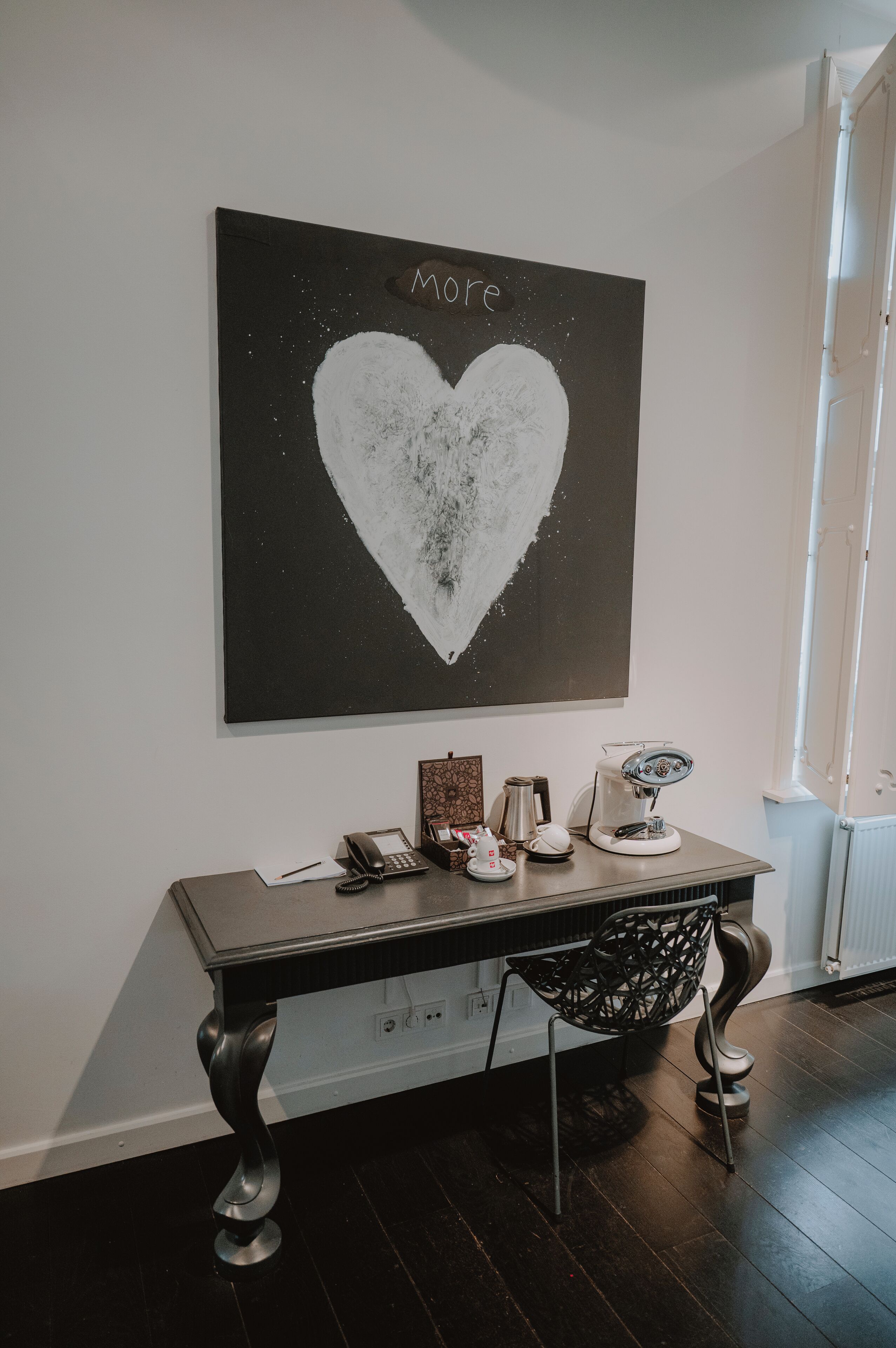Una habitación con un escritorio de madera oscura, un teléfono blanco, una cafetera y un objeto decorativo sobre el escritorio. En la pared, encima del escritorio, hay una obra de arte en blanco y negro con forma de corazón.