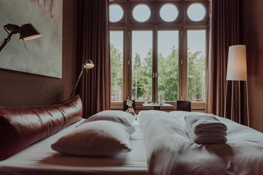 A hotel room with a bed, a lamp, a painting, and a view of trees through large windows.