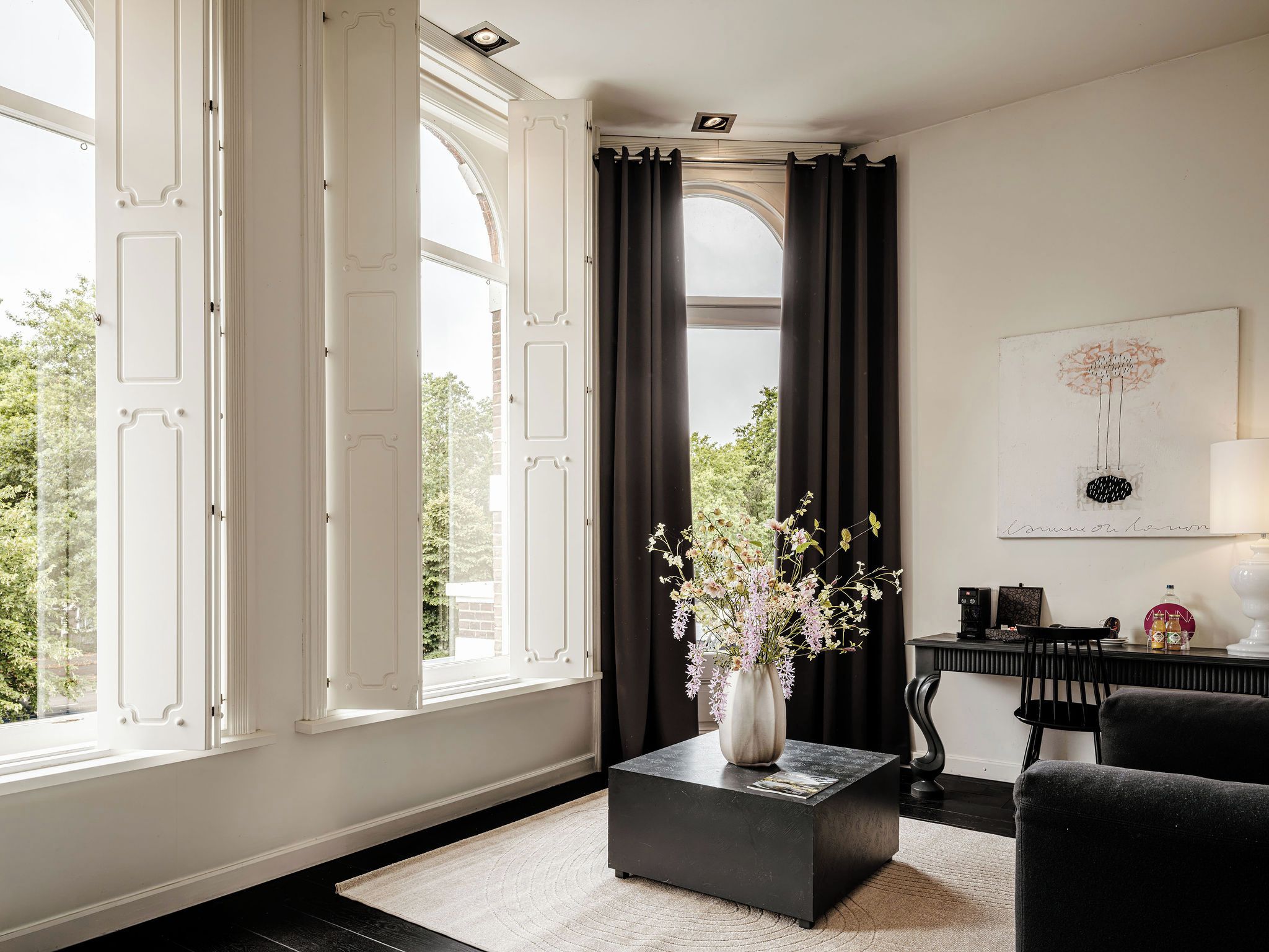 A room with a large window, a black coffee table, a chair, and a painting on the wall.