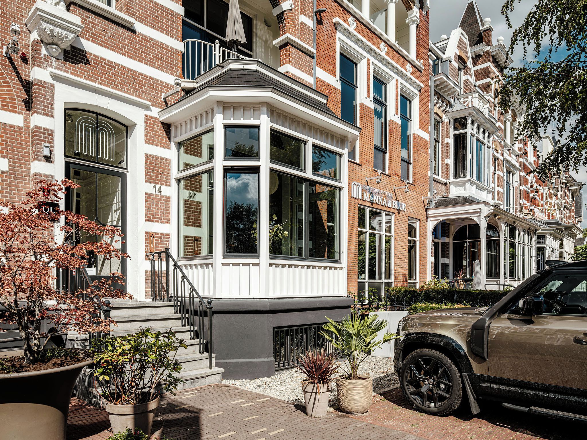 Een rij herenhuisachtige woningen met erkers, een bord met de naam van een boetiekhotel, en een geparkeerde SUV.