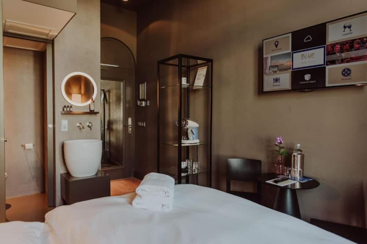 Habitación de hotel con una cama, un espejo redondo, una mesita con un florero y una pantalla en la pared con varios iconos.