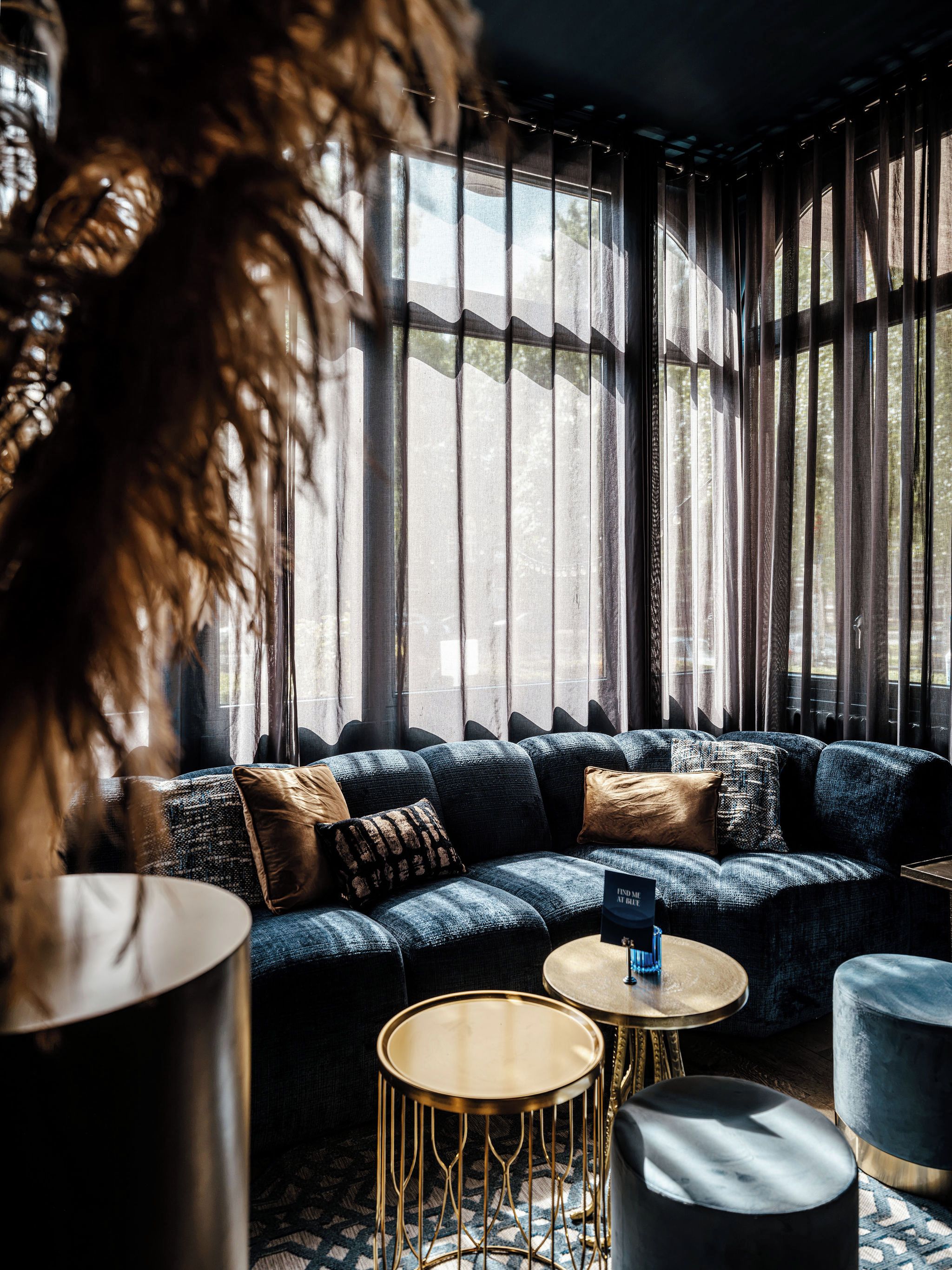 A modern lounge area with a blue sofa, round gold tables, and a large window with curtains.