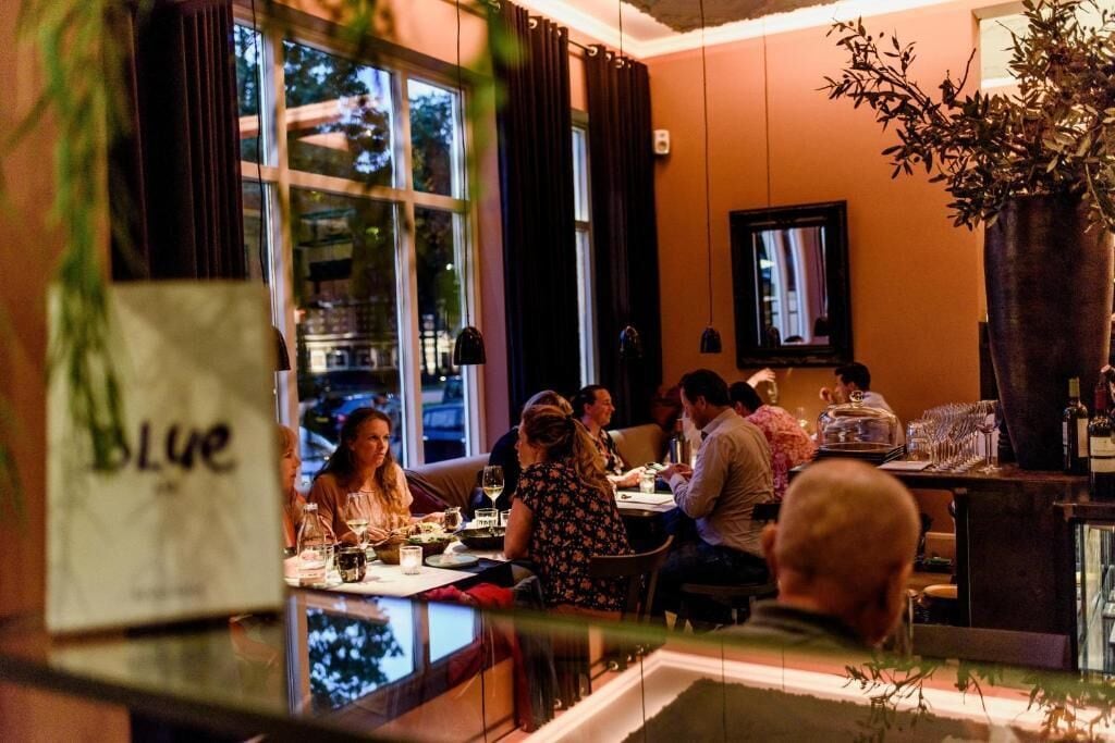Un restaurante con comensales en las mesas, ventanas amplias y una planta en maceta.