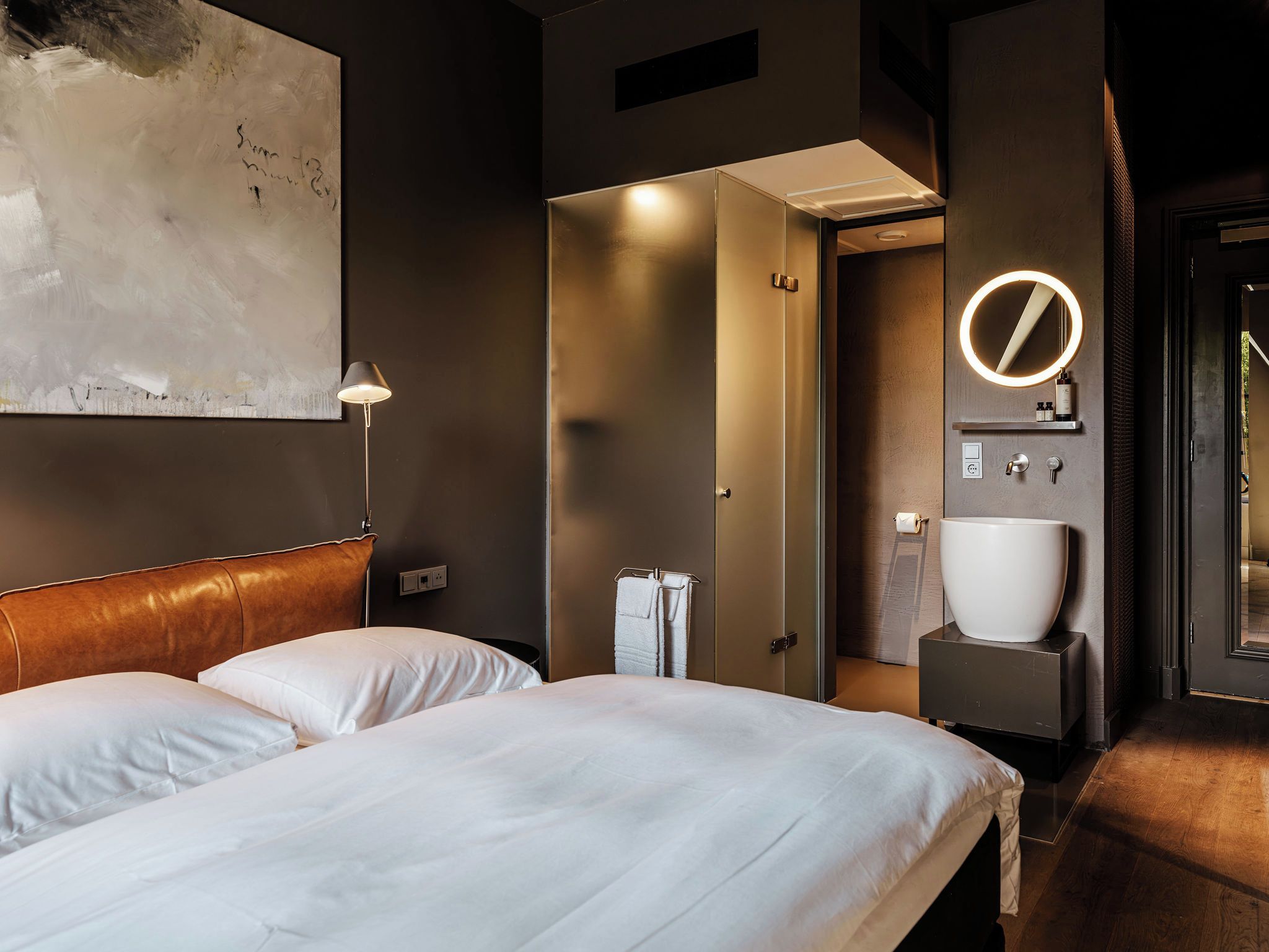A hotel room with a bed, a bedside lamp, a bathroom with a sink and mirror, and a door leading to another room.