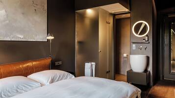 A hotel room with a bed, a bedside lamp, a bathroom with a sink and mirror, and a door leading to another room.