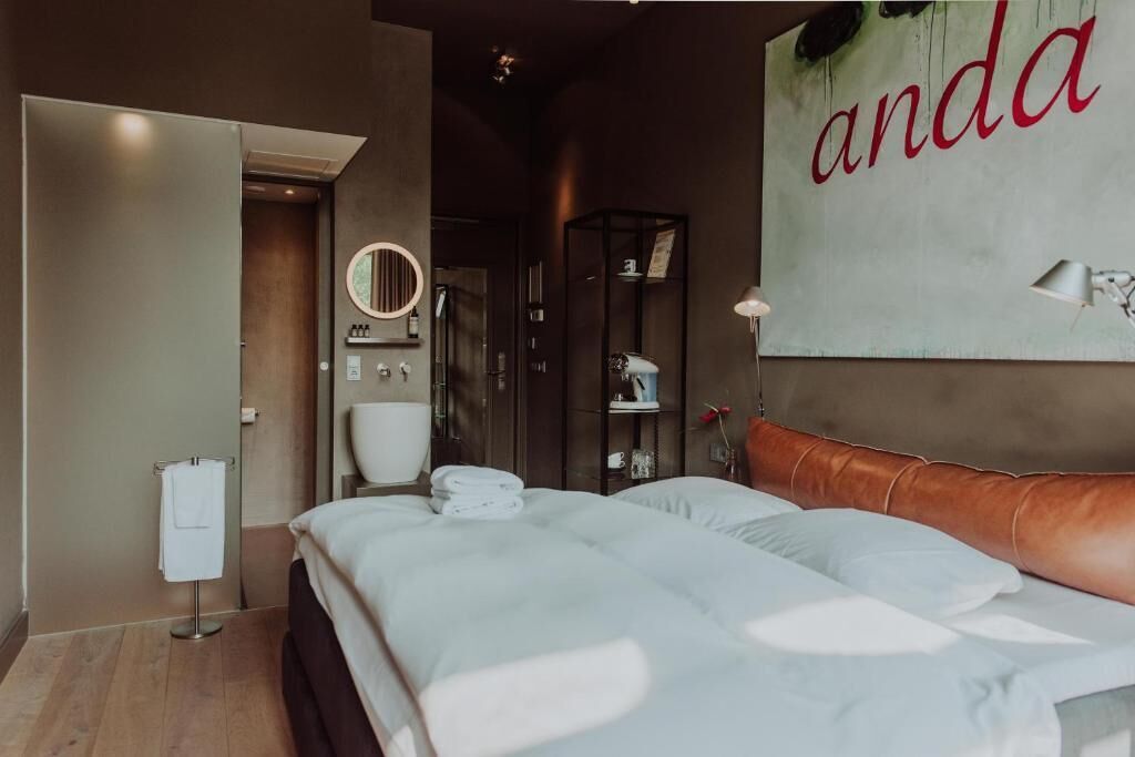 A modern hotel room with a bed, a bathroom with a toilet and sink, a mirror, and a wall with the word 'anda' in red letters.