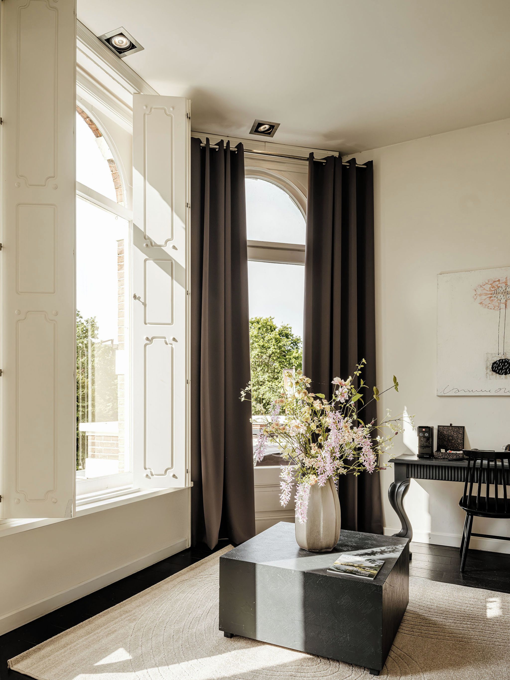 A room with a large window, a black coffee table with a vase of flowers, a black chair, and a desk with a computer.