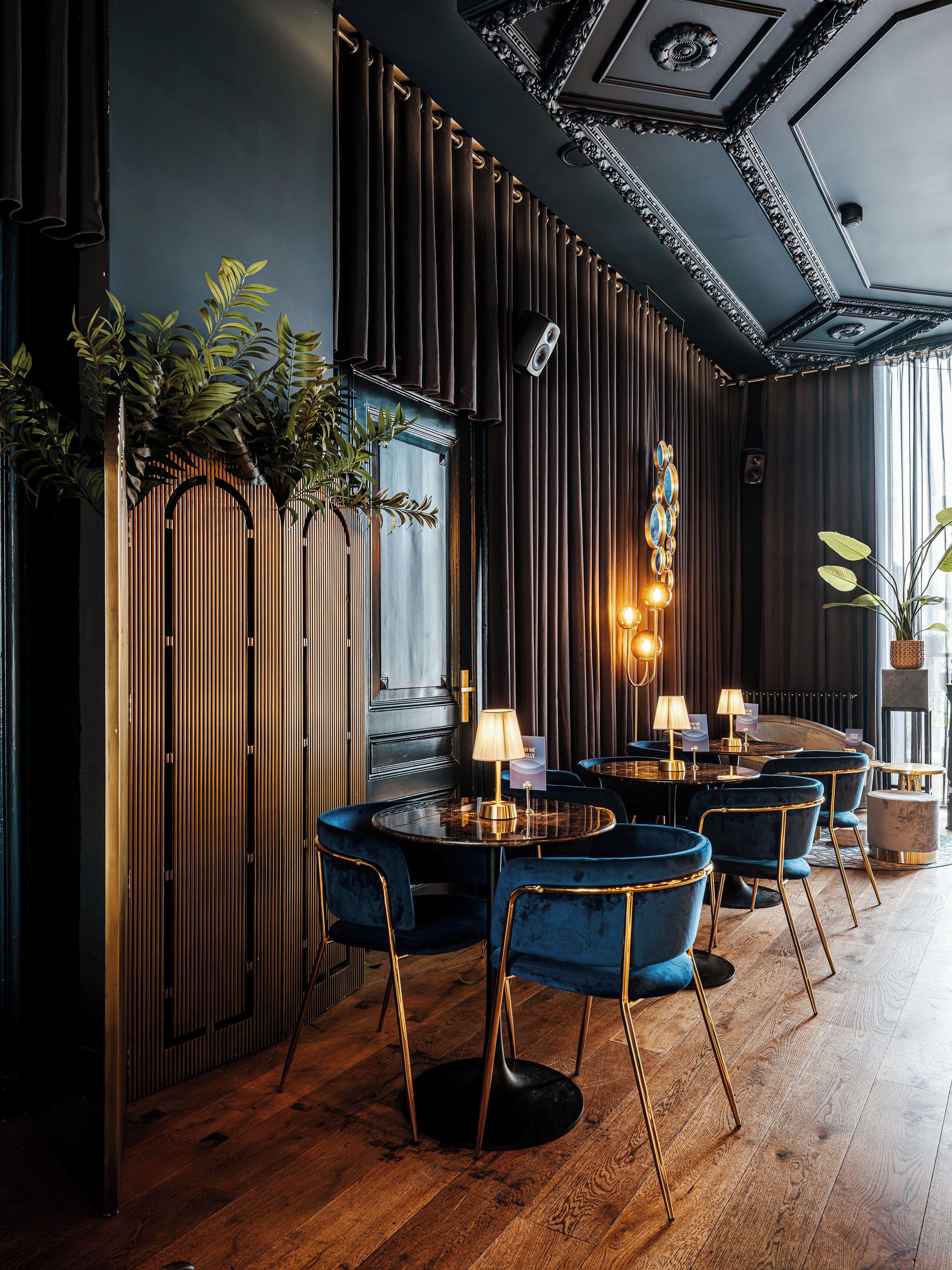 Interior de restaurante con suelo de madera, mesas redondas con sillas y paneles de madera verticales.