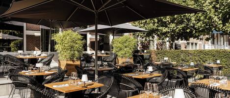 Un área de comedor al aire libre con varias mesas y sillas, sombrillas y plantas en macetas.