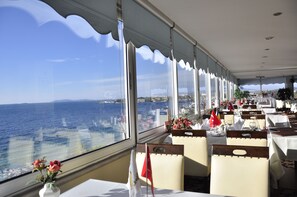 A restaurant with tables set for dining, large windows offering a view of the sea, and flags on the tables. - Otel Yelkenkaya (Darica)
