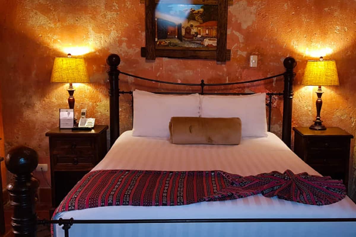A bed with a white sheet and a patterned blanket, flanked by two nightstands with lamps, in a room with a rustic brick wall and a painting above the bed.