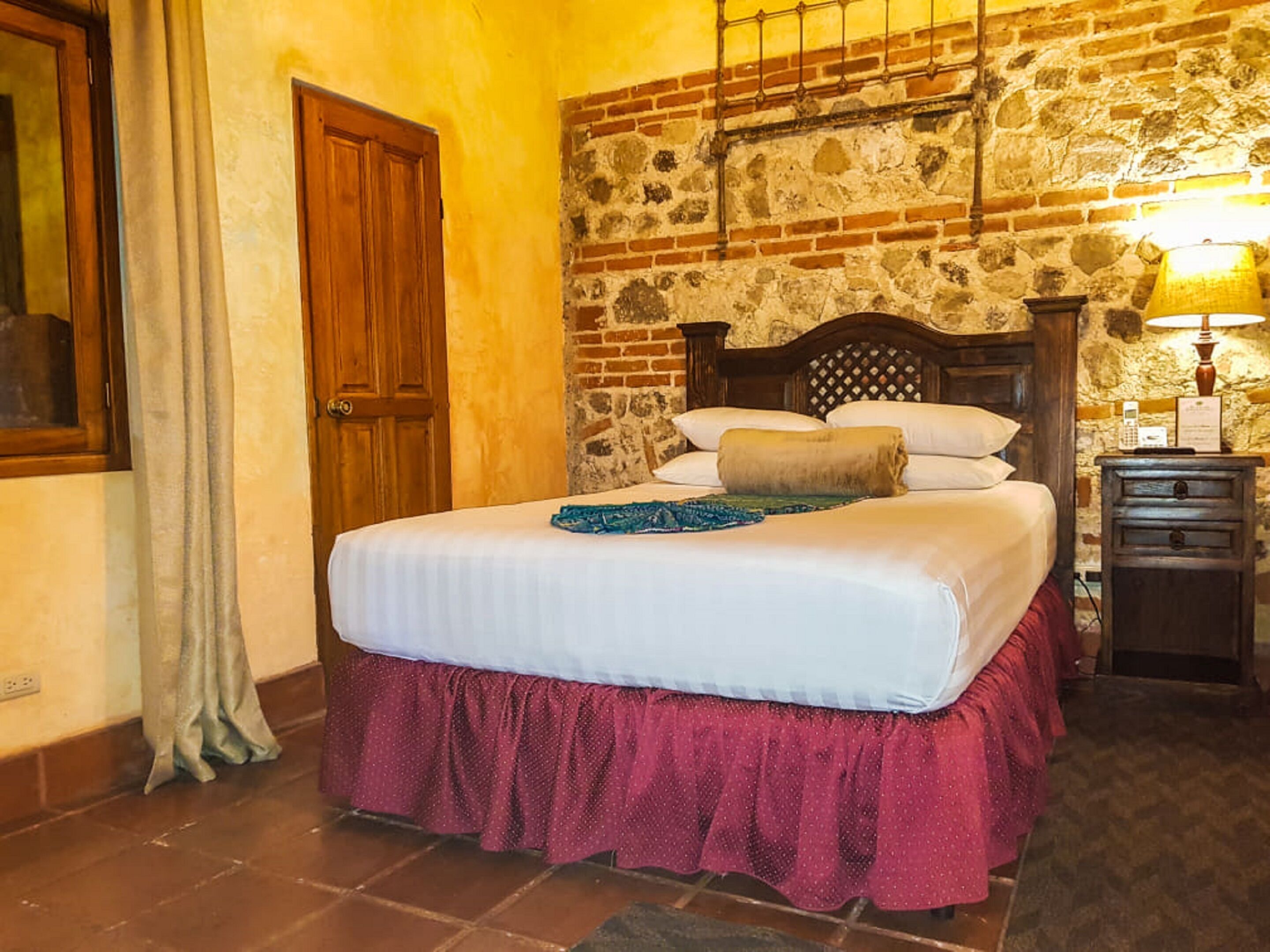 A hotel room with a bed, a wooden headboard, a brick wall, a wooden nightstand, and a lamp.