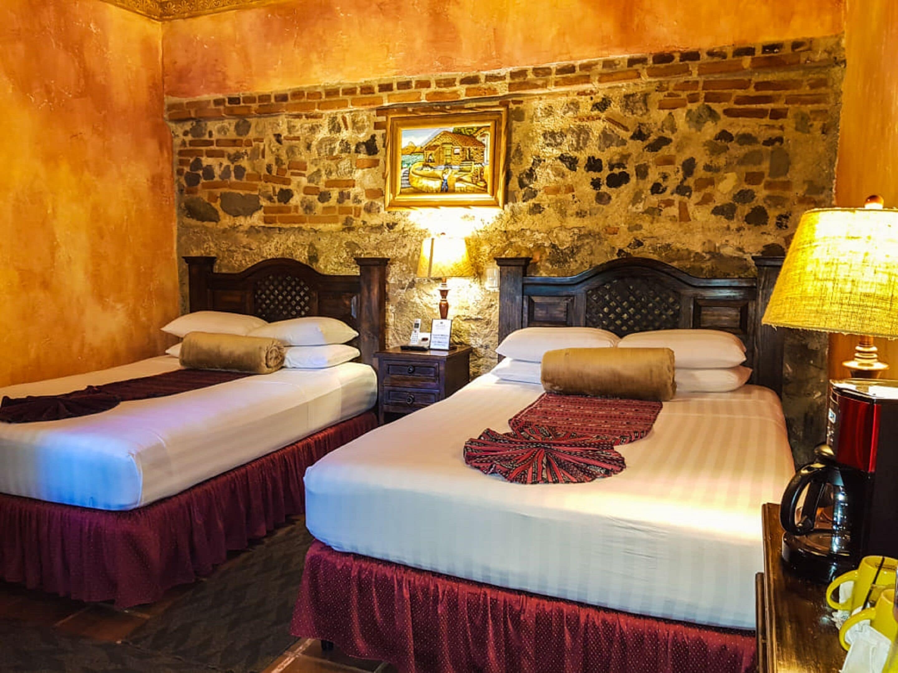 Habitación con dos camas, cada una con cabecero, una mesita de noche con lámpara y una pared de piedra con un cuadro enmarcado.