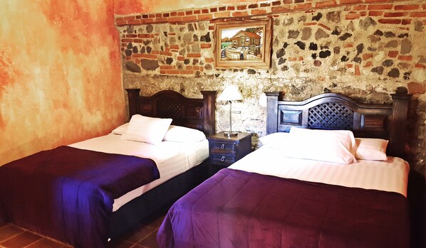 Una habitación con dos camas, una mesita de noche con lámpara y una pared de piedra con un cuadro enmarcado.
