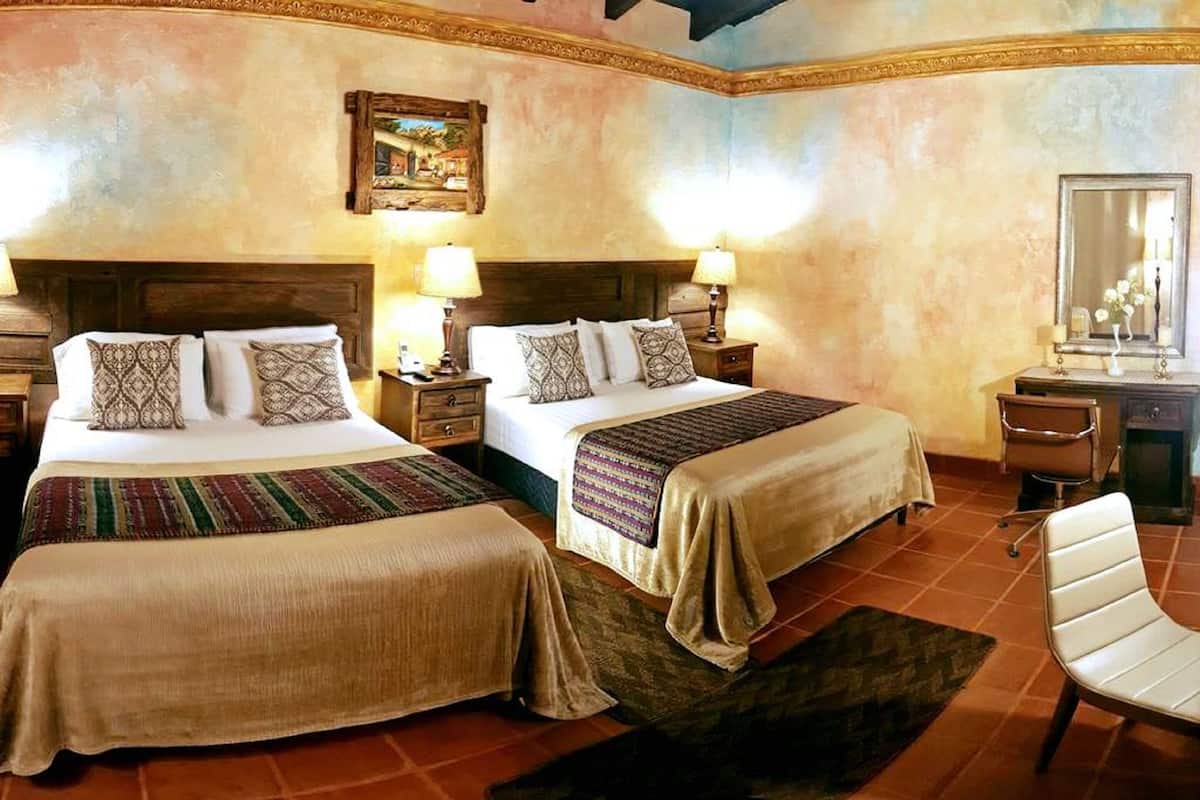 A hotel room with two beds, each with a wooden headboard and matching nightstands. There is a desk with a chair and a mirror.