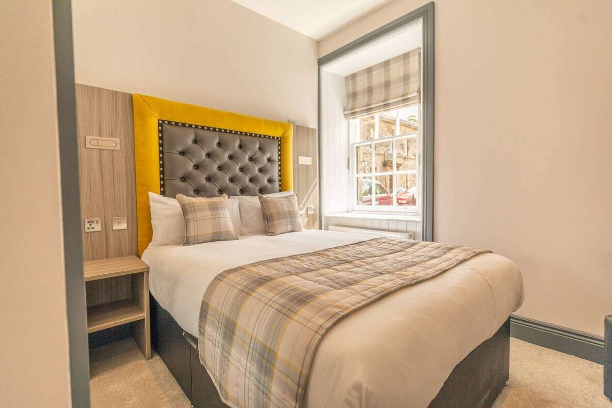 Une chambre à coucher avec une tête de lit capitonnée, un lit recouvert d’une couverture à carreaux, une fenêtre donnant sur l’extérieur et une armoire en bois.